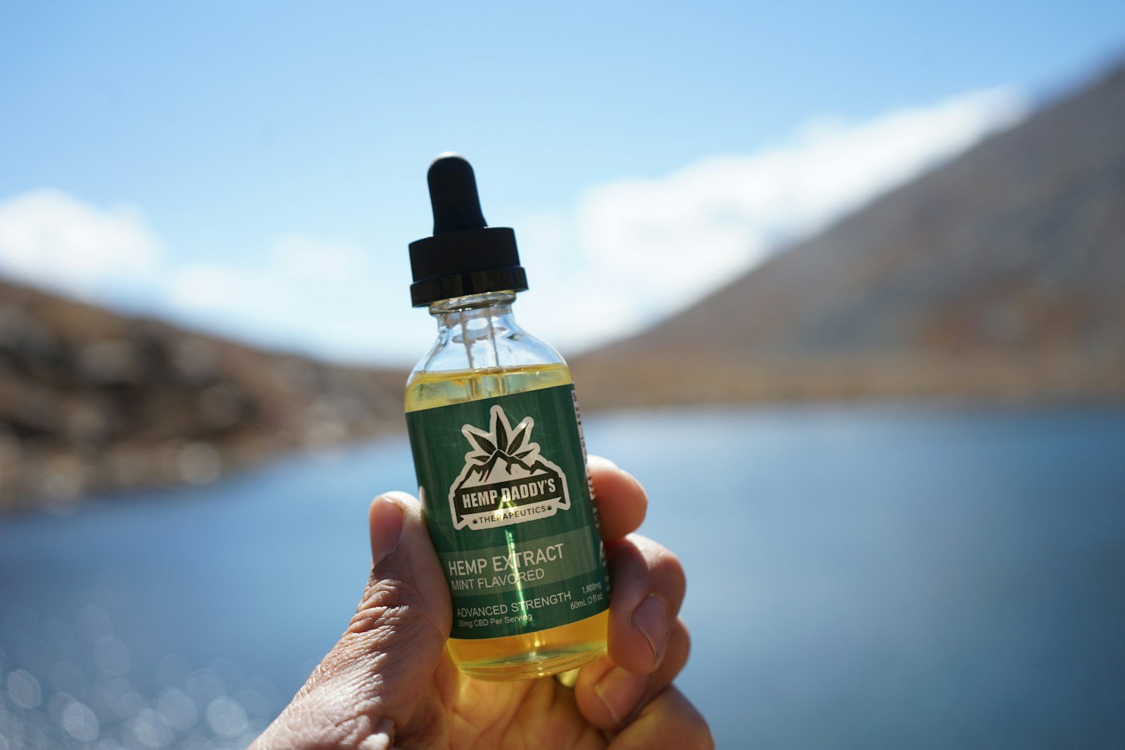 Hand holding Hemp Daddy’s hemp extract oil bottle outdoors near a lake