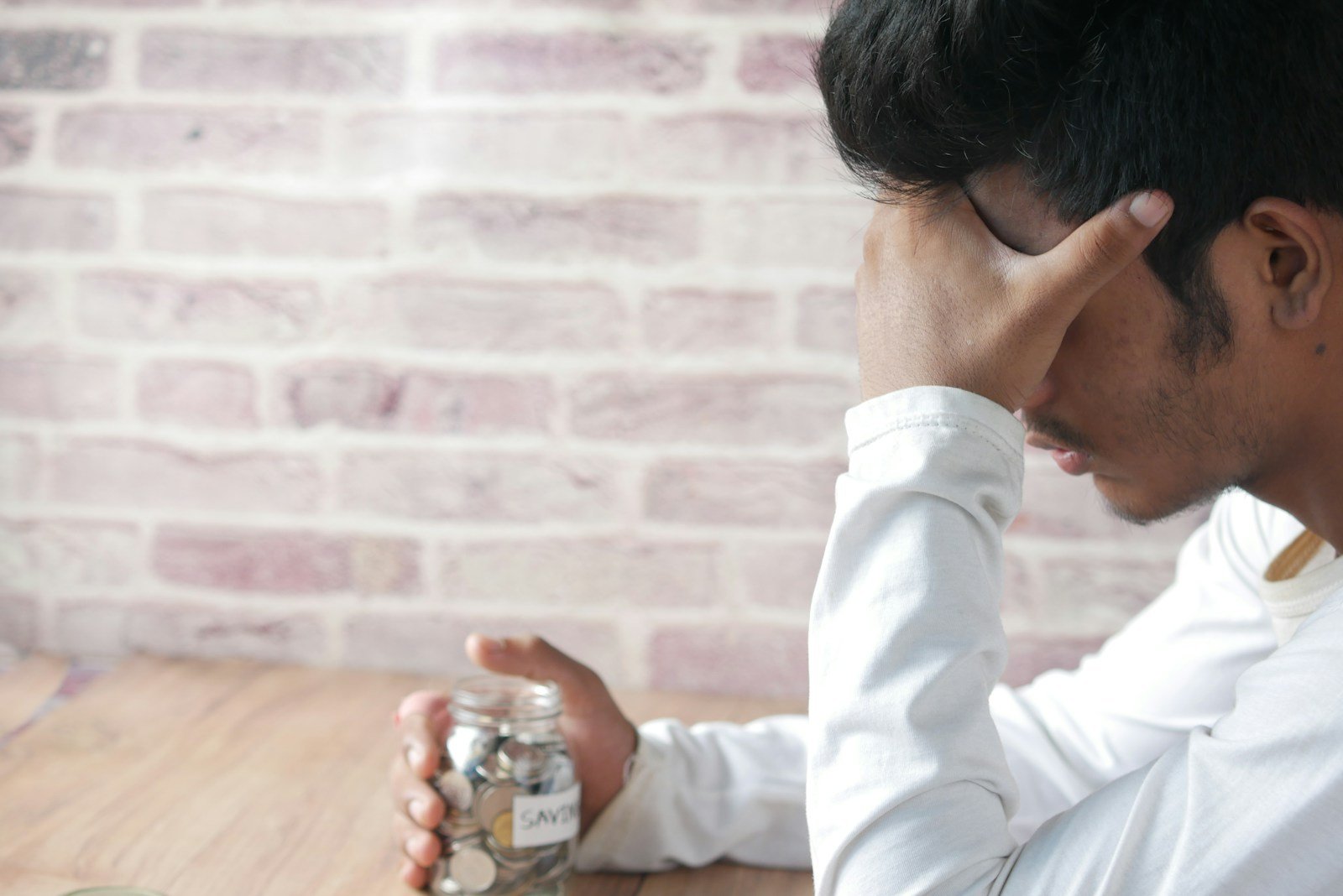 Stressed man holding head with hand while clutching a small savings jar of coins