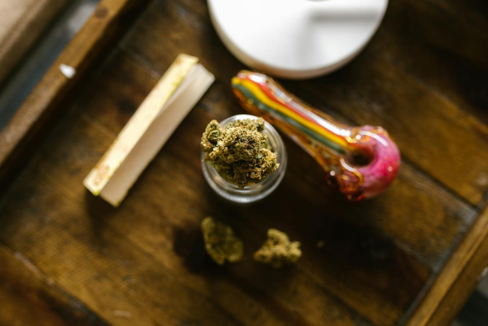 Jar of cannabis buds with rolling papers and glass smoking pipe on wooden tray
