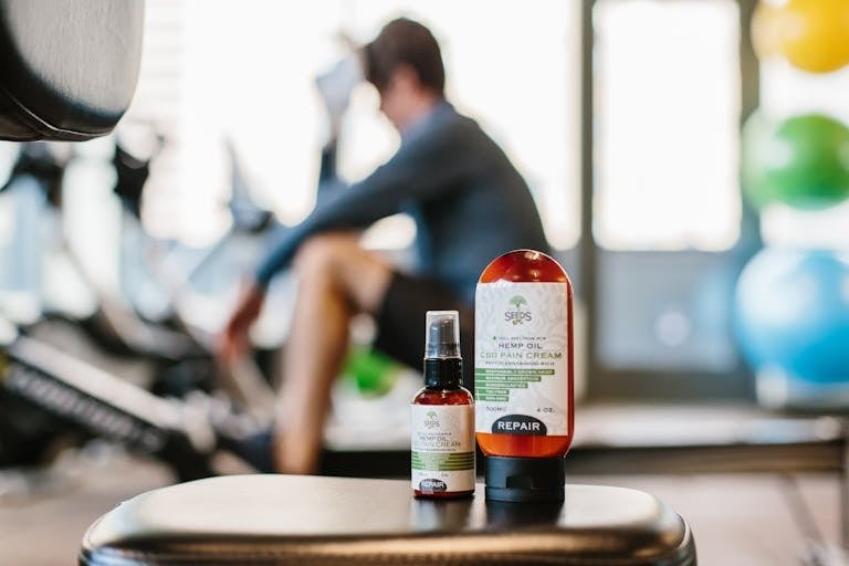 CBD hemp oil pain relief cream bottles with athlete resting in gym background